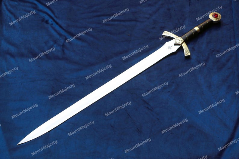 May include: A silver sword with a gold hilt and a red jewel on the pommel. The sword is lying on a blue fabric background.