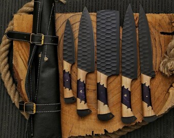 May include: A set of six black chef knives with wooden handles and purple accents, displayed on a wooden cutting board. The blades have a textured surface. A black leather knife roll is also present.