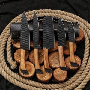 May include: Set of six black knives with wooden handles, arranged on a pile of wooden rounds and surrounded by a thick rope. The knives have a textured, honeycomb-like pattern on the blades.