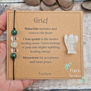 May include: A brown card with the word "Grief" in large letters. The card has text about the healing properties of malachite, clear quartz, and moonstone. The card also has a small white angel figurine and a bracelet with green and white beads.