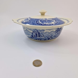 Könnte beinhalten: Eine blau-weiße Keramikschale mit Deckel. Die Schale zeigt ein Landschaftsdesign mit Gebäuden, Bäumen und Figuren. Der Deckel hat ein dekoratives Muster und einen kleinen Knopf. Die Schale hat einen Durchmesser von etwa 15 cm.