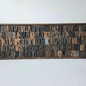 May include: A wooden printer's tray filled with individual wooden letterpress blocks. The blocks are a mix of letters, numbers, and symbols in various fonts and sizes. The tray is rectangular and made of dark wood.