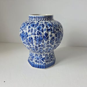 May include: A blue and white ceramic vase with a floral pattern. The vase has a bulbous body and a flared base. The rim is decorated with a repeating swirl design. The vase is a decorative item.