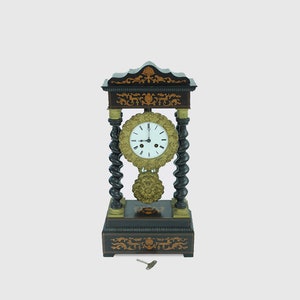 Puede incluir: Un reloj antiguo negro y dorado con una esfera blanca y marcadores de hora en números romanos. El reloj tiene un diseño decorativo con detalles intrincados y una llave en la base.