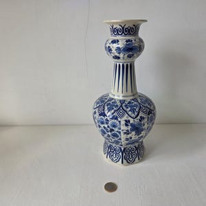 May include: An elegant blue and white ceramic vase with a distinctive shape. The vase has a rounded base, a slender neck, and a flared top, adorned with floral patterns and vertical stripes. The vase is approximately 30 cm tall.