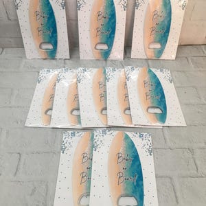 May include: A collection of surfboard-shaped bottle openers. Each opener is painted with a gradient of blue and peach, with the words "Baby on Board" in black script. They are packaged on white cards with blue confetti.