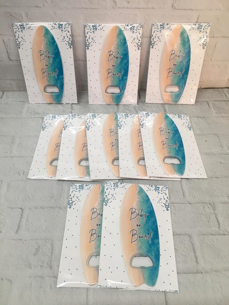 May include: A collection of surfboard-shaped bottle openers. Each opener is painted with a gradient of blue and peach, with the words "Baby on Board" in black script. They are packaged on white cards with blue confetti.