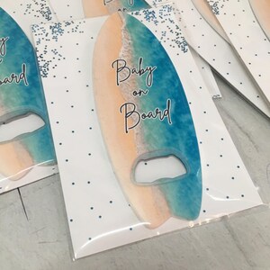 May include: Surfboard-shaped bottle opener with a beach-themed design. The opener features a gradient of colors, including blue, tan, and white, with the words "Baby on Board" in elegant script. The opener is packaged on a white card with blue polka dots.