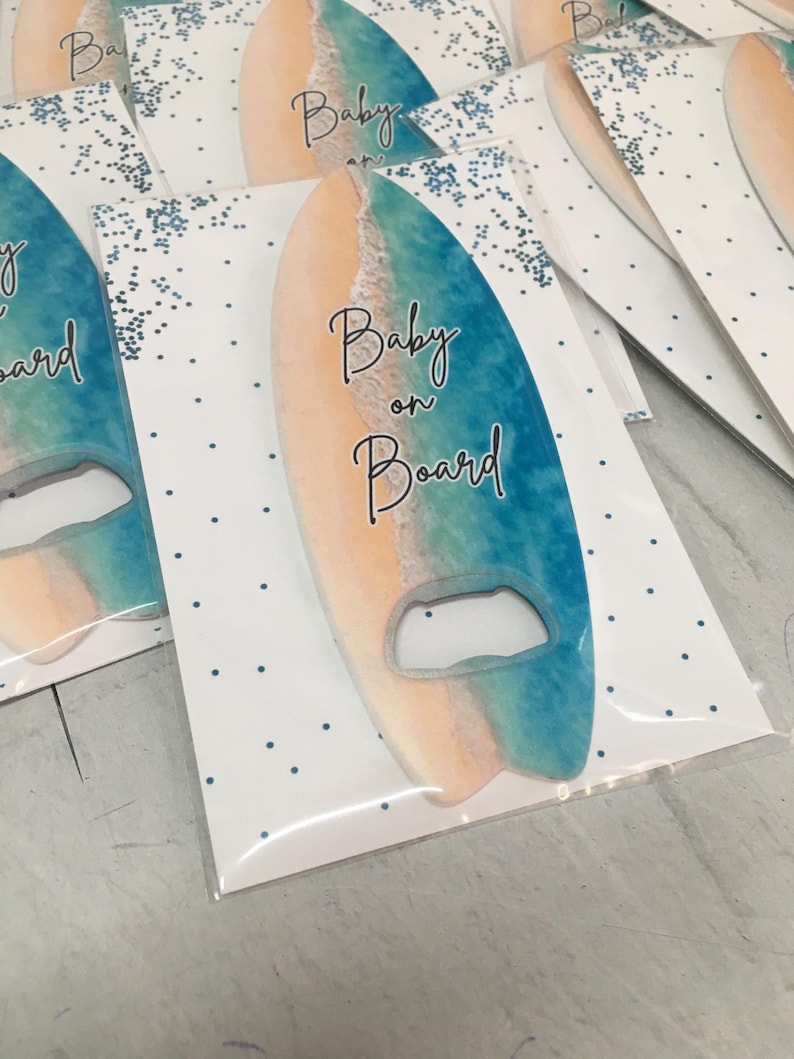 May include: Surfboard-shaped bottle opener with a beach-themed design. The opener features a gradient of colors, including blue, tan, and white, with the words "Baby on Board" in elegant script. The opener is packaged on a white card with blue polka dots.