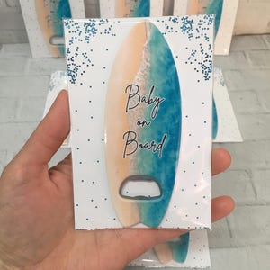 May include: A surfboard-shaped bottle opener with a beach scene design, featuring tan, blue, and white colors. The words "Baby on Board" are written on the front. Several packaged openers are visible.