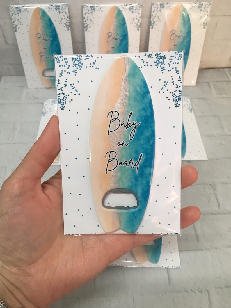 May include: A surfboard-shaped bottle opener with a beach scene design, featuring tan, blue, and white colors. The words "Baby on Board" are written on the front. Several packaged openers are visible.