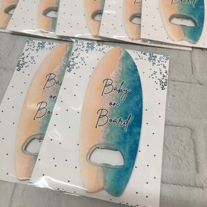 May include: Surfboard-shaped bottle openers with a beach-themed design. The openers feature a gradient of colors, including tan, white, and blue, with the words "Baby on Board" in elegant script. The openers are packaged on white cards with blue dot accents.