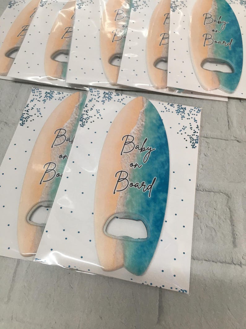 May include: Surfboard-shaped bottle openers with a beach-themed design. The openers feature a gradient of colors, including tan, white, and blue, with the words "Baby on Board" in elegant script. The openers are packaged on white cards with blue dot accents.