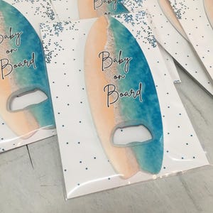 May include: Surfboard-shaped bottle opener with a beach scene design. The opener is blue and tan, with the words "Baby on Board" in black script. The opener is packaged in a clear plastic sleeve with a white card featuring blue polka dots and glitter.