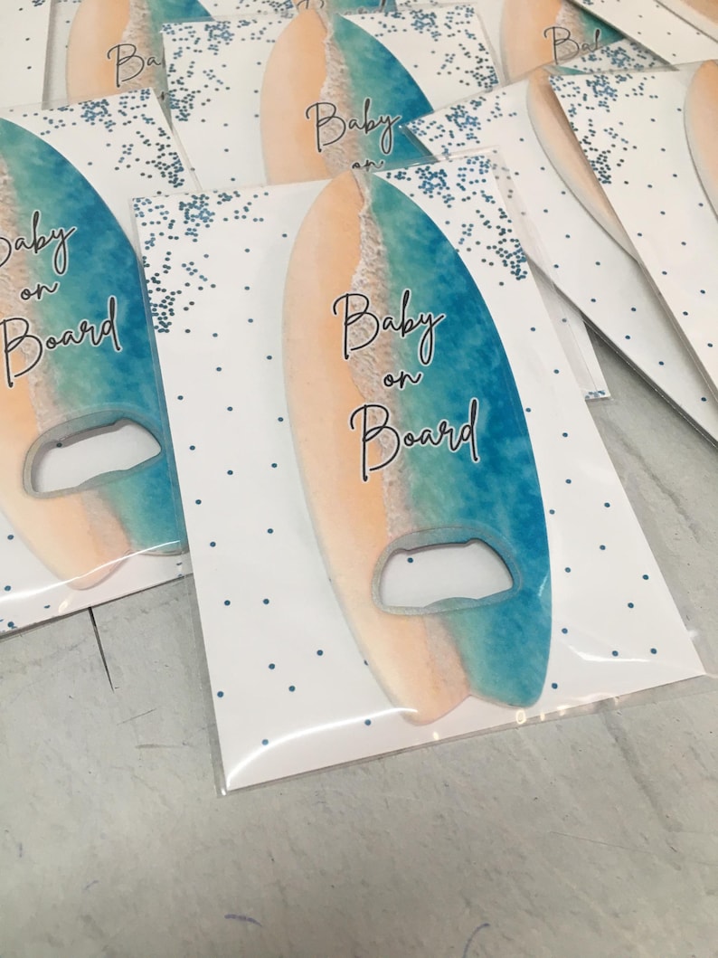 May include: Surfboard-shaped bottle opener with a beach scene design. The opener is blue and tan, with the words "Baby on Board" in black script. The opener is packaged in a clear plastic sleeve with a white card featuring blue polka dots and glitter.