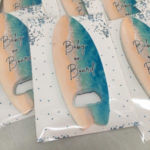 May include: Surfboard-shaped bottle openers with a beach-themed design. The openers are in a clear plastic packaging with the words "Baby on Board" printed on them. The surfboard design features a gradient of colors, including blue, beige, and white.
