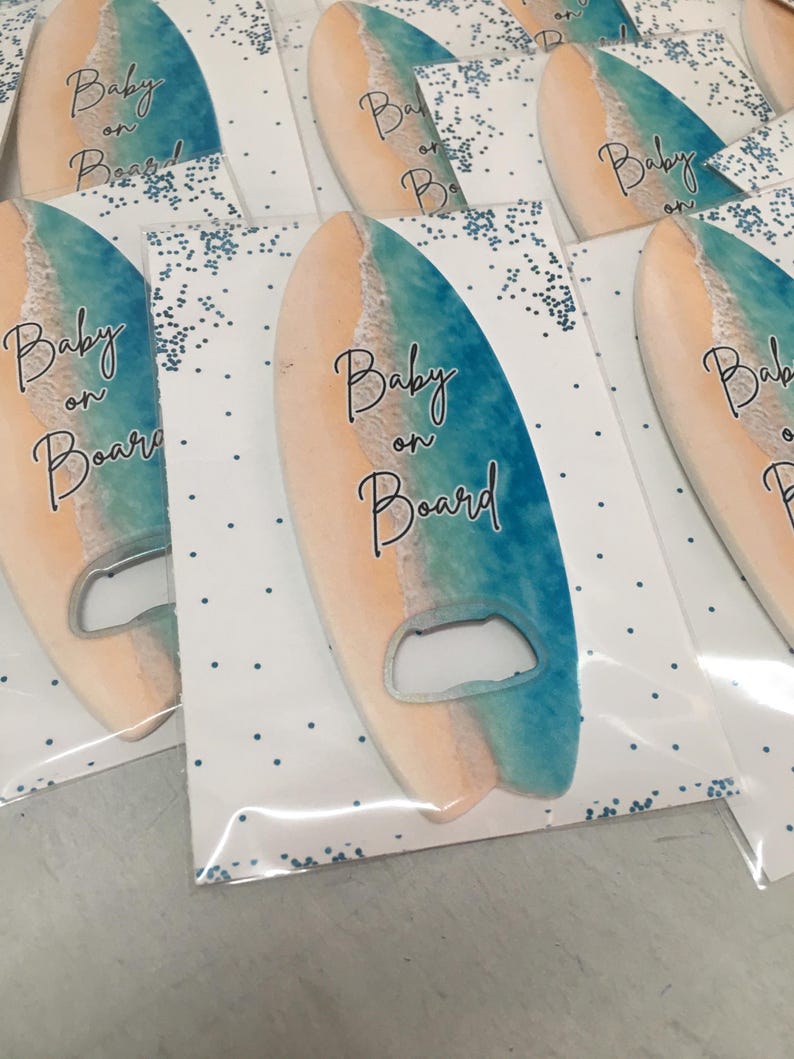 May include: Surfboard-shaped bottle openers with a beach-themed design. The openers are in a clear plastic packaging with the words "Baby on Board" printed on them. The surfboard design features a gradient of colors, including blue, beige, and white.