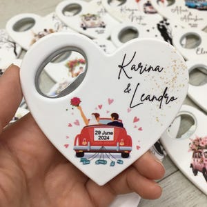 May include: White heart-shaped bottle opener with a silver metal opener. The design features a red car with a bride and groom, the date "29 June 2024", and the names "Karina & Leandro". Other openers are visible in the background.