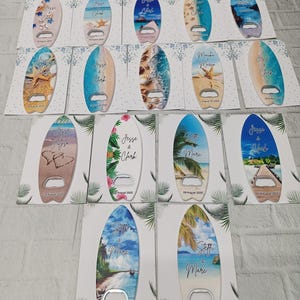 May include: A collection of surfboard-shaped bottle openers with beach-themed designs. Each opener features a unique ocean scene, including starfish, shells, and tropical landscapes. The names "Jesse & Clark" and "Stoffi & Mare" are printed on some openers, along with the date "09 August 2025".