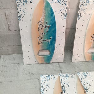 May include: Surfboard-shaped bottle opener with a beach-themed design. The opener features a gradient of colors, including tan, white, and blue, with the words "Baby on Board" in elegant script. The opener is mounted on a white card with blue confetti.
