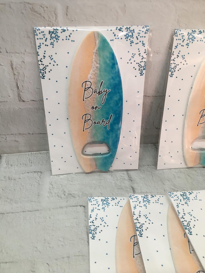 May include: Surfboard-shaped bottle opener with a beach-themed design. The opener features a gradient of colors, including tan, white, and blue, with the words "Baby on Board" in elegant script. The opener is mounted on a white card with blue confetti.