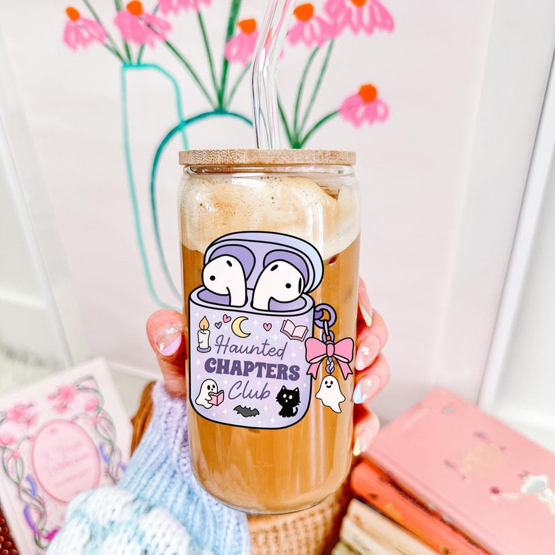 A cute spooky bookish png tumbler cup mockup of a haunted themed earbuds case for readers who love audiobooks. The case has the text "Haunted Chapers Club". The case is decorated with a pink bow, ghosts, sparkles, a bat, black cat, candle, book and the moon.