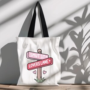 May include: White canvas tote bag with a pink and red sign that says "Smut St" and "Lovers Lane" with hearts. The sign is on a wooden post with green grass at the bottom.
