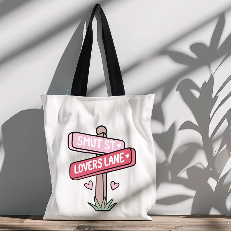 May include: White canvas tote bag with a pink and red sign that says "Smut St" and "Lovers Lane" with hearts. The sign is on a wooden post with green grass at the bottom.