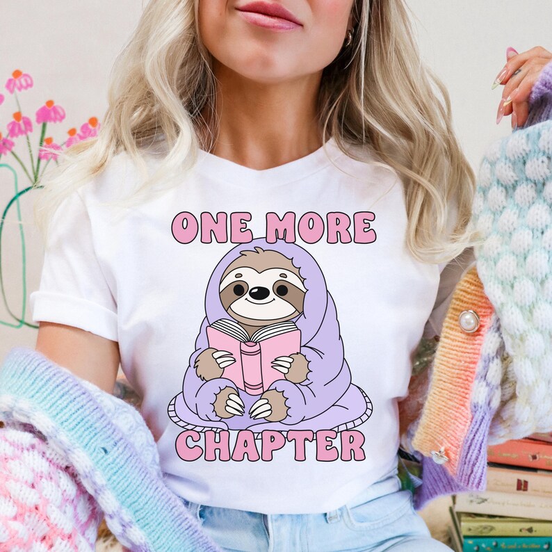 A white t-shirt showing the one more chapter png design. A cozy sloth reading a book.