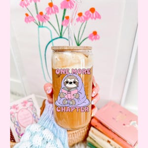 A glass can cup showing the one more chapter png design. A cozy sloth reading a book.
