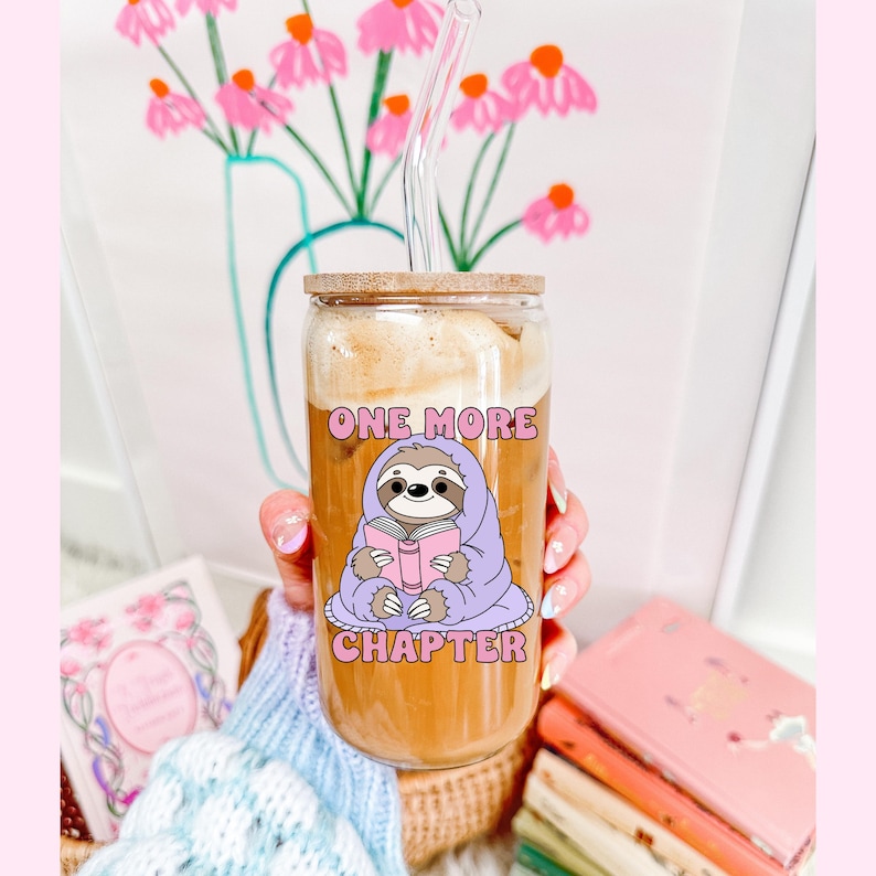 A glass can cup showing the one more chapter png design. A cozy sloth reading a book.