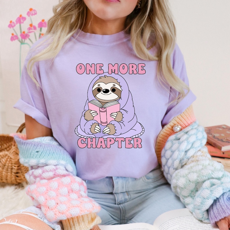 A purple t-shirt showing the one more chapter png design. A cozy sloth reading a book.