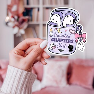 A Halloween bookish png sticker mockup of a girly hand holding the the png design. The design is of a haunted themed headphones case for readers who love audiobooks. The case has the text "Haunted Chapers Club". The case is decorated with a pink bow, ghosts, sparkles, a bat, black cat, candle, book and the moon.