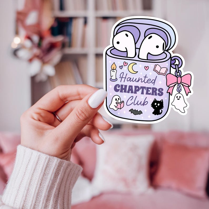 A Halloween bookish png sticker mockup of a girly hand holding the the png design. The design is of a haunted themed headphones case for readers who love audiobooks. The case has the text "Haunted Chapers Club". The case is decorated with a pink bow, ghosts, sparkles, a bat, black cat, candle, book and the moon.
