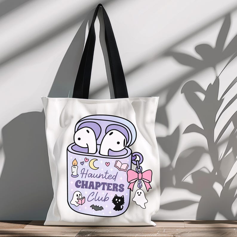 A cute Halloween bookish png tote bag mockup of a haunted themed earbuds case for readers who love audiobooks. The case has the text "Haunted Chapers Club". The case is decorated with a pink bow, ghosts, sparkles, a bat, black cat, candle, book and the moon.