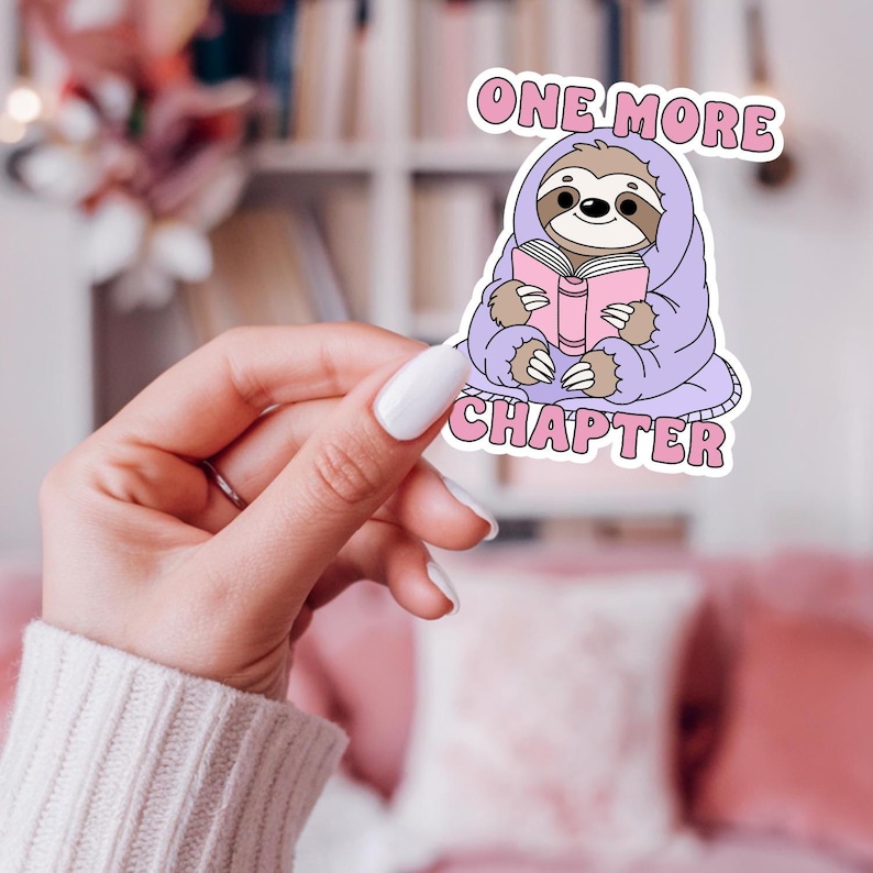 A sticker mockup of the One More Chapter Bookish PNG. A pretty female hand holds up the design in front of a bookshelf.