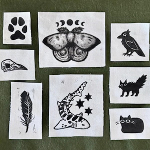 Può includere: Una collezione di otto toppe in tessuto rettangolari con disegni serigrafati in inchiostro nero su sfondo bianco. I motivi includono un'impronta di zampa, una falena con fasi lunari, un teschio di uccello, una piuma, uno squalo con stelle, un corvo, un gatto e un gatto seduto.