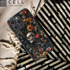 May include: A black phone case with a floral and skull design. The case features a dark background with colorful flowers, leaves, and a white skull. The phone case has a cutout for the camera lens.