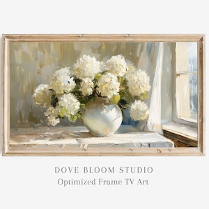 Pode incluir: Uma pintura emoldurada de um buquê de hortênsias brancas em um vaso branco sobre uma mesa em frente a uma janela. A pintura está em uma moldura de madeira clara.