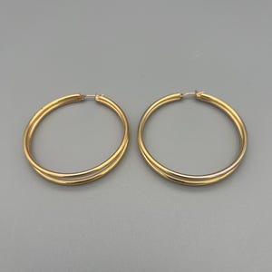 Könnte beinhalten: Zwei goldfarbene Creolen. Jeder Ohrring hat ein großes, kreisförmiges Design mit glatter, polierter Oberfläche. Die Ohrringe bestehen aus glänzendem Metall und werden vor grauem Hintergrund präsentiert.