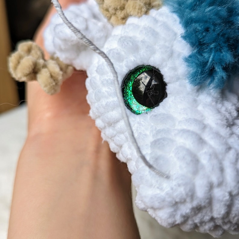May include: A white crocheted stuffed animal with a green glitter eye. The animal has a blue and brown fur accent.