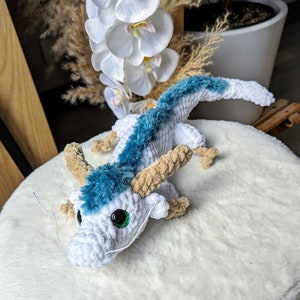 May include: A white crocheted dragon with blue fur and green eyes. The dragon is sitting on a white fluffy surface.