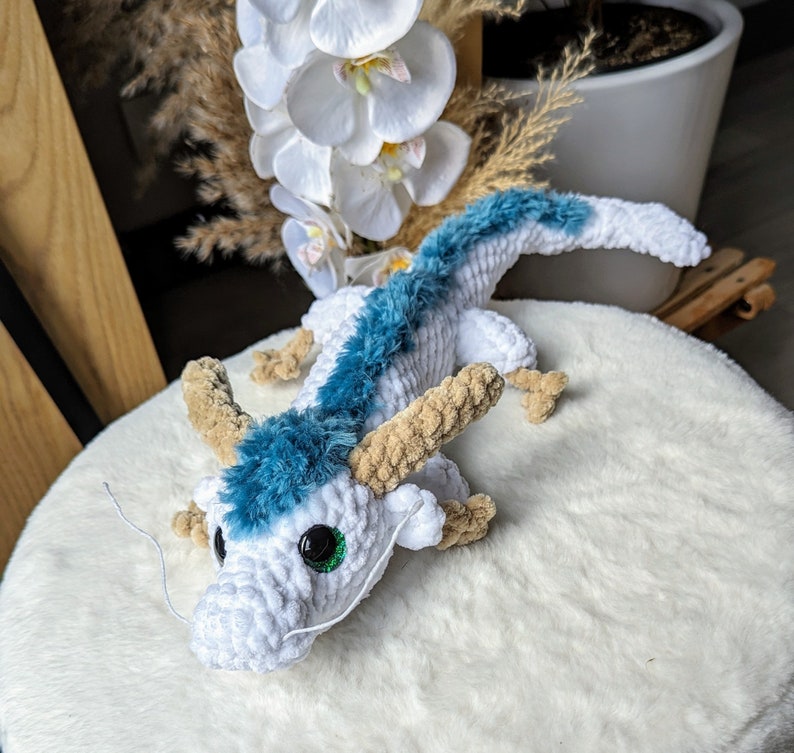 May include: A white crocheted dragon with blue fur and green eyes. The dragon is sitting on a white fluffy surface.