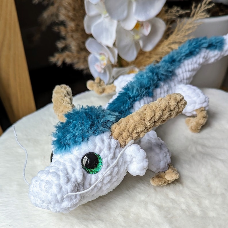 May include: A white and blue crocheted dragon toy with a green eye. The dragon has a long, curled tail and a small, pointed snout.