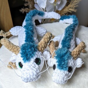 May include: Two white crocheted dragon plushies with blue fur accents and green eyes. The dragons are lying on a white surface.