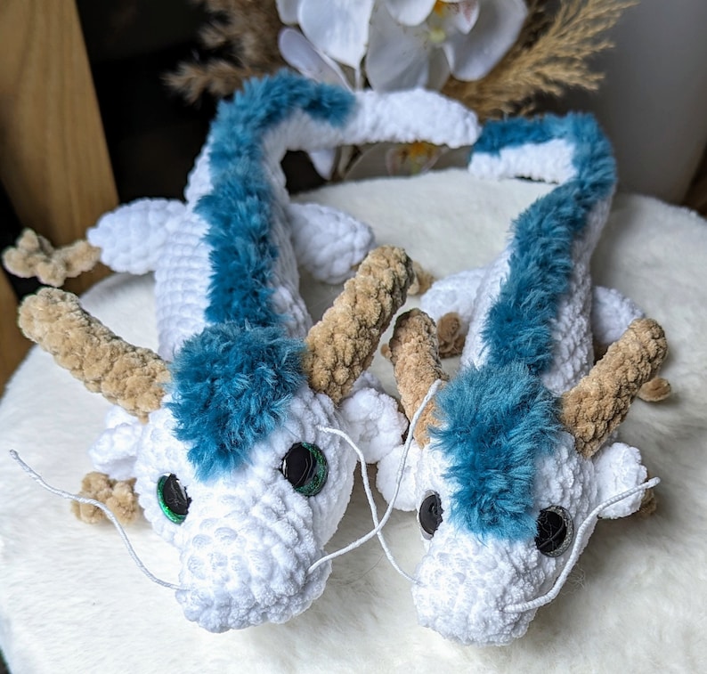 May include: Two white crocheted dragon plushies with blue fur accents and green eyes. The dragons are lying on a white surface.