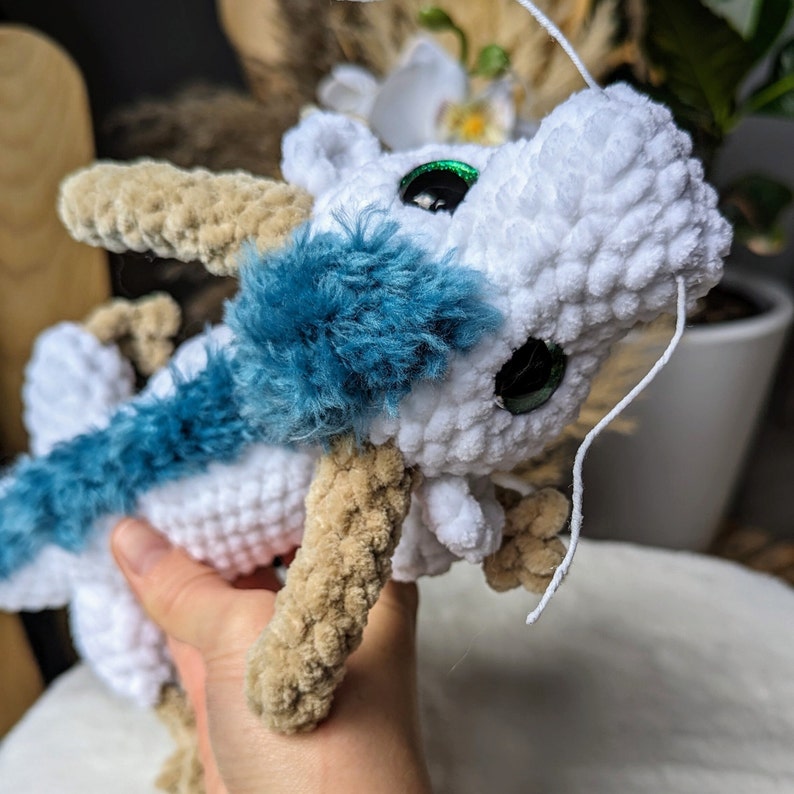 May include: A white crocheted dragon with blue fur accents and green eyes. The dragon has a long, thin tail and is holding its head up.