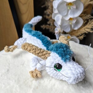 May include: A white crocheted dragon with blue fur accents and green eyes. The dragon is lying on a white surface.