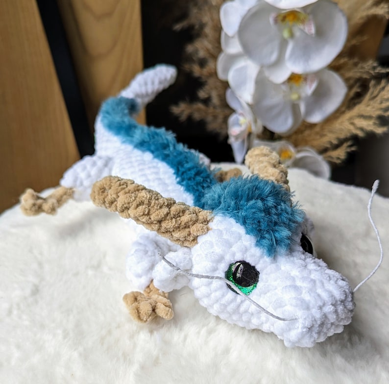 May include: A white crocheted dragon with blue fur accents and green eyes. The dragon is lying on a white surface.
