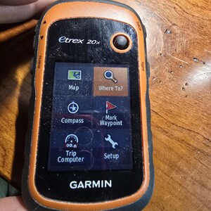 May include: A black and orange Garmin etrex 20x handheld GPS device with a color screen displaying a map, compass, and other navigation features. The screen also shows the text "Where To?" and "Mark Waypoint".